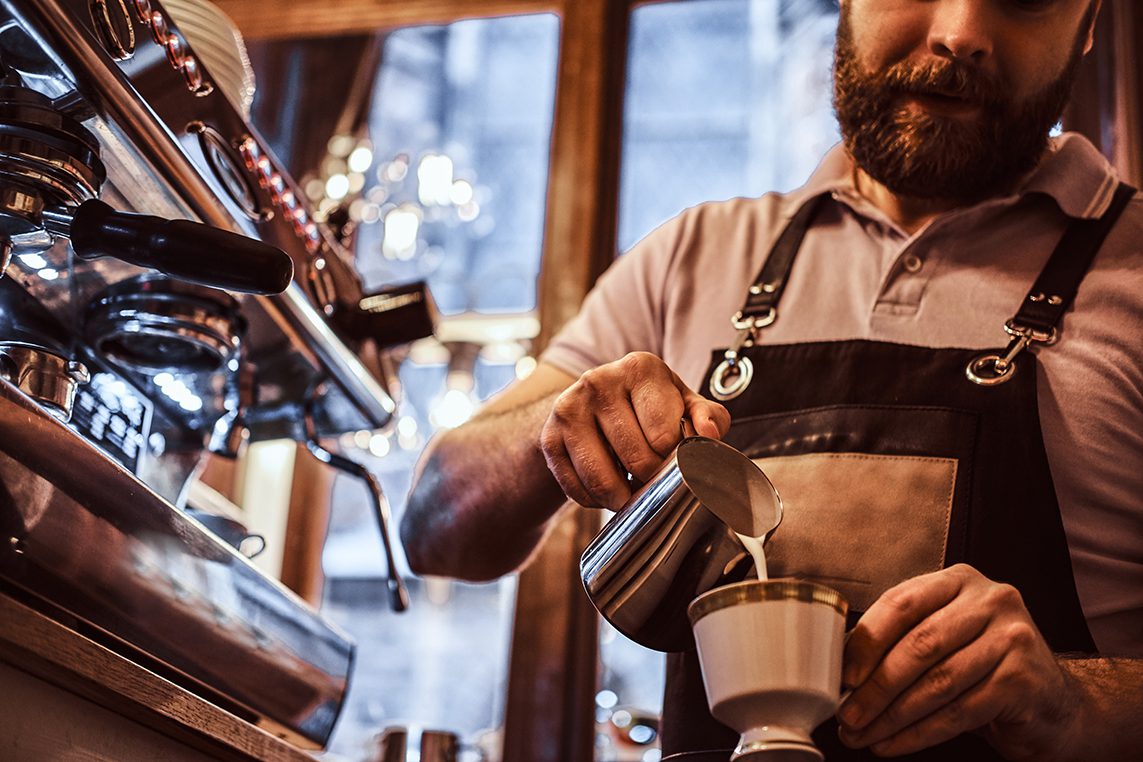 Barista wearing apron making a cappuccino, pouring milk in a cup in a restaurant or coffee shop photo of barista